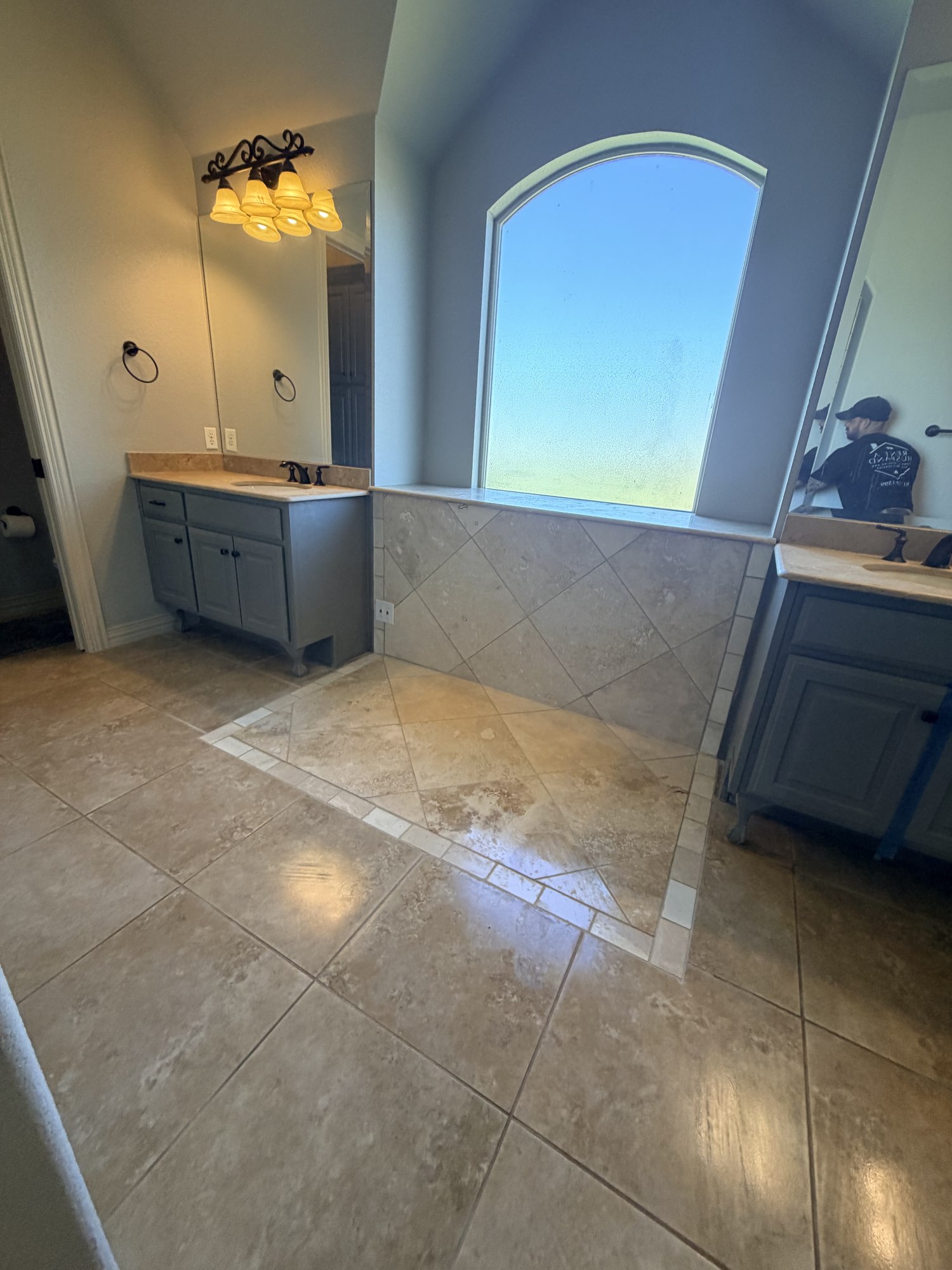 Bathroom with gray vanities and a tiled floor and wall beneath an arched window.
